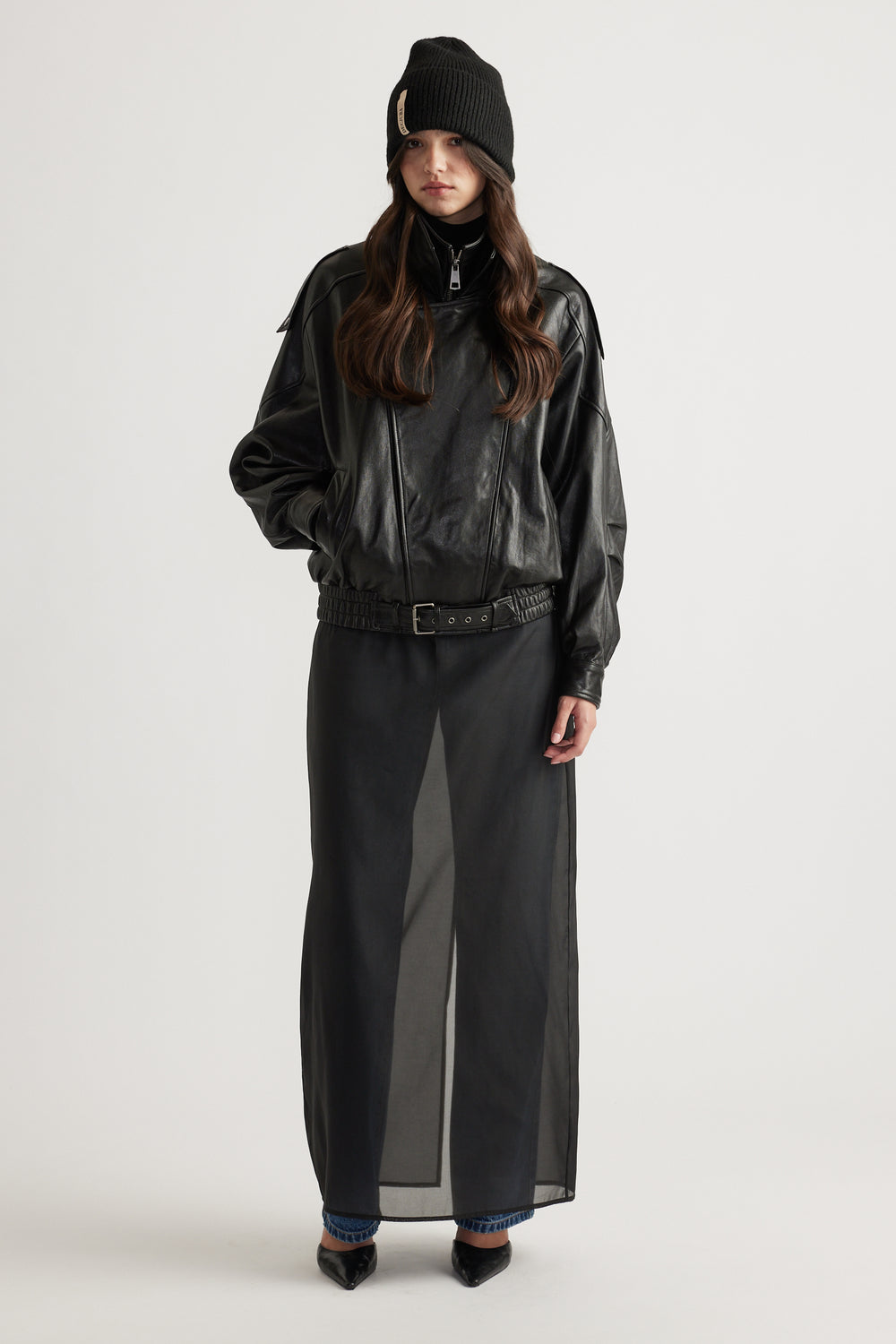 Model wearing a black knit cuff beanie paired with a loose-fit black zip-up jacket and a sheer black maxi skirt.
