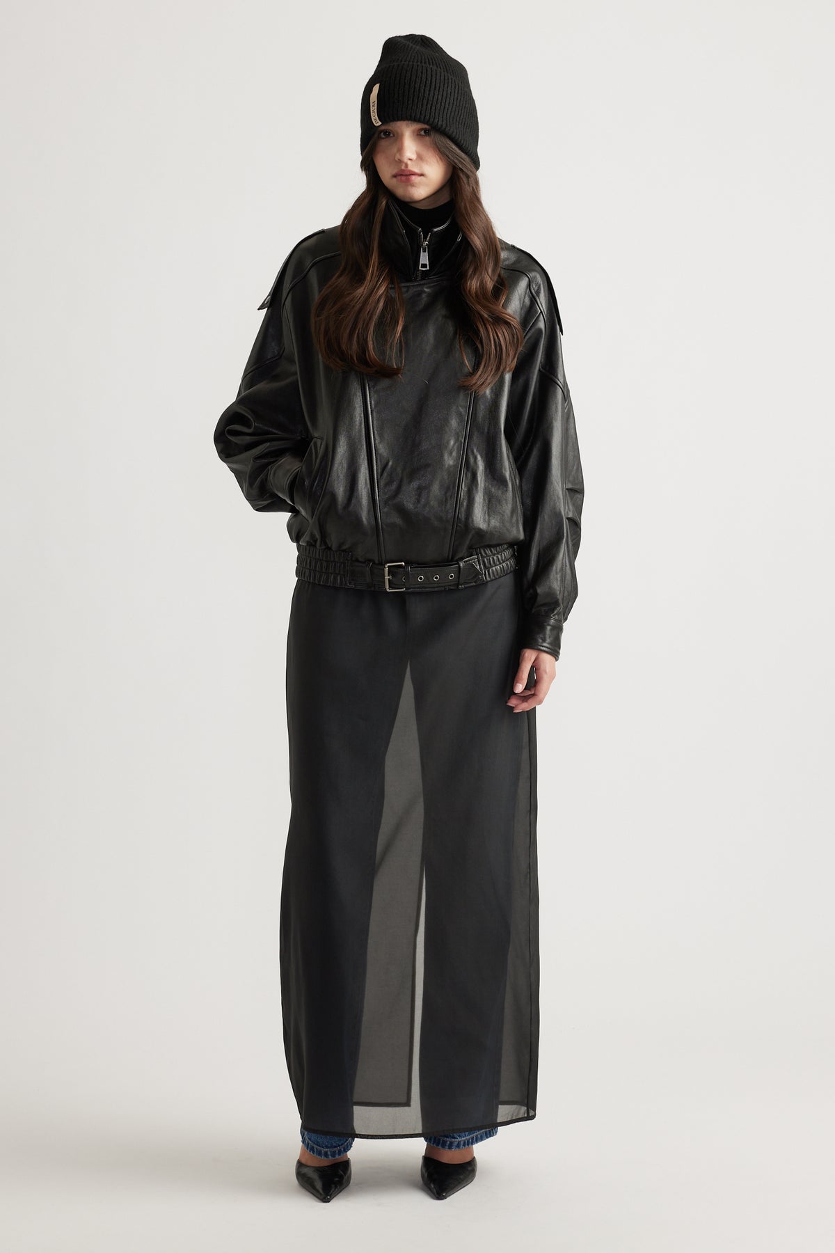 Model wearing a black knit cuff beanie paired with a loose-fit black zip-up jacket and a sheer black maxi skirt.