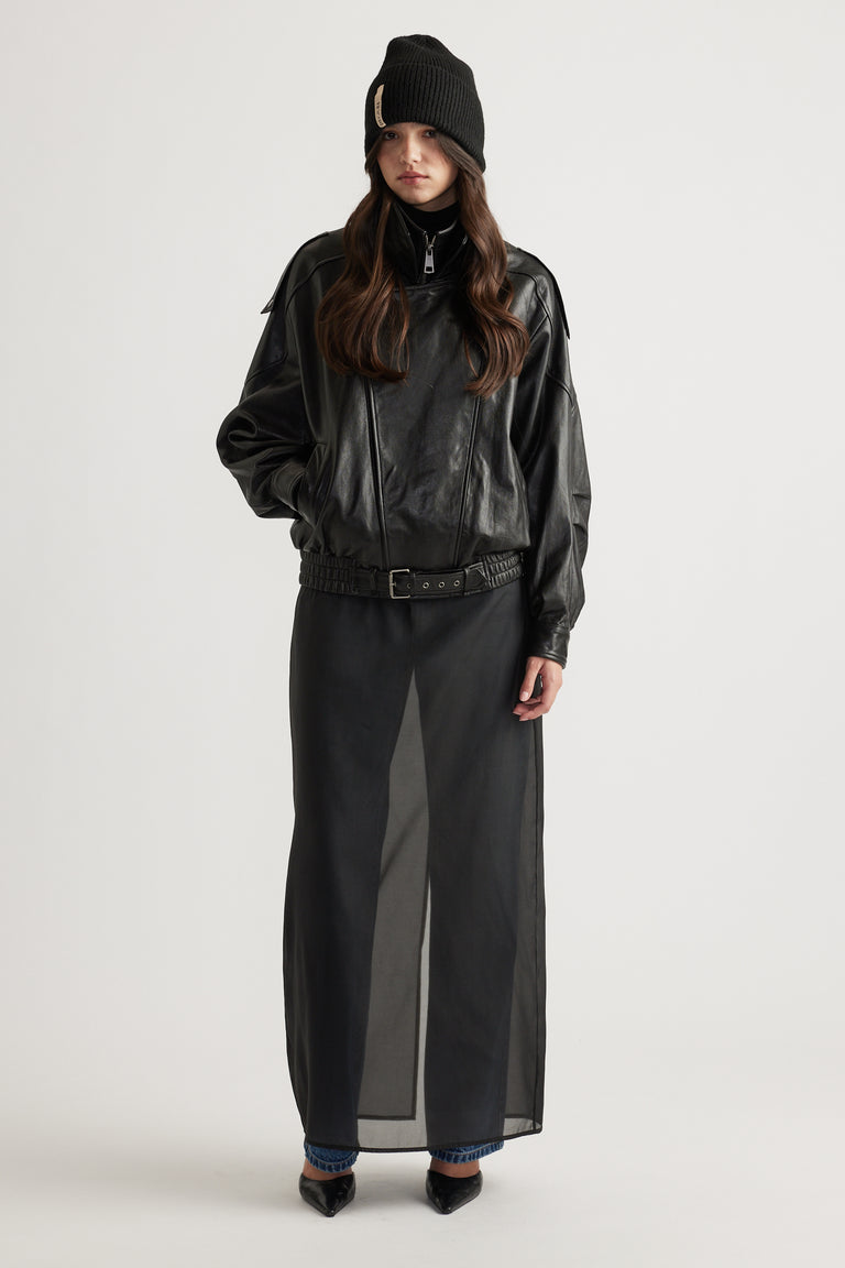Model wearing a black knit cuff beanie paired with a loose-fit black zip-up jacket and a sheer black maxi skirt.