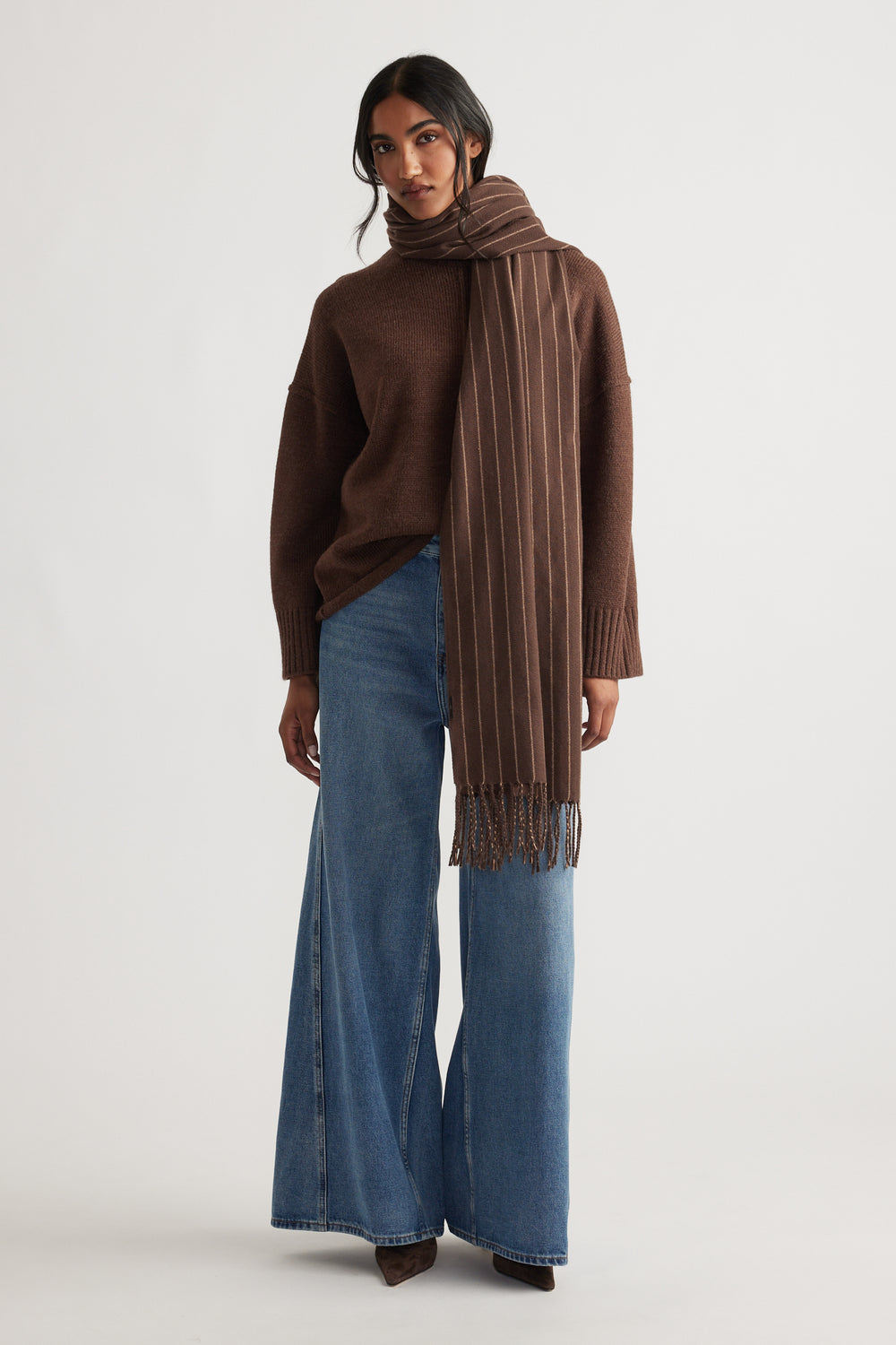 Woman wearing Gia Stripe Scarf in warm brown tones paired with wide-leg denim jeans and a cozy knit sweater.