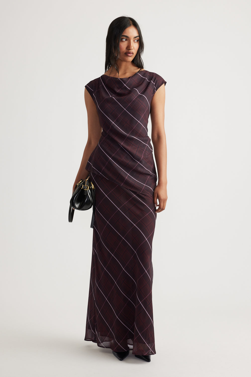 Woman wearing a sleeveless dark plaid maxi dress with a draped neckline, holding a small black handbag.
