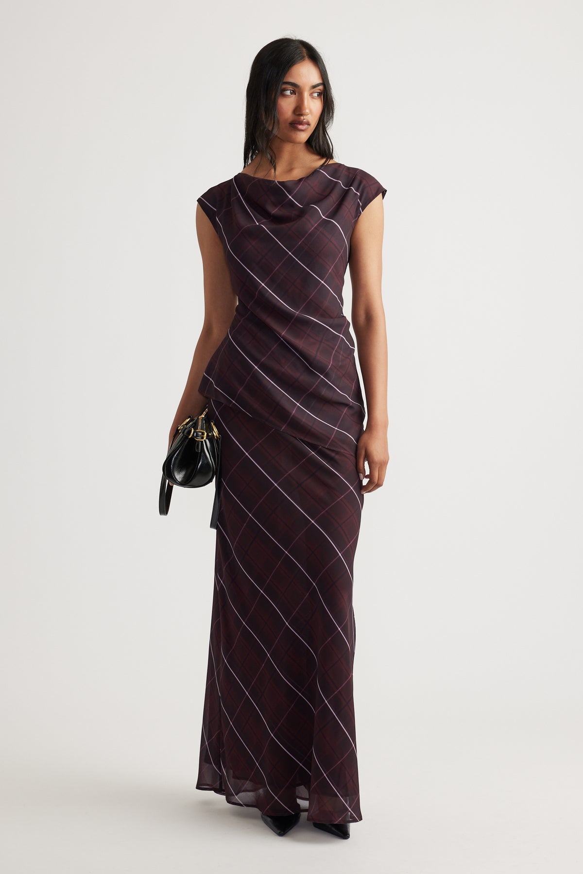 Woman wearing a sleeveless dark plaid maxi dress with a draped neckline, holding a small black handbag.