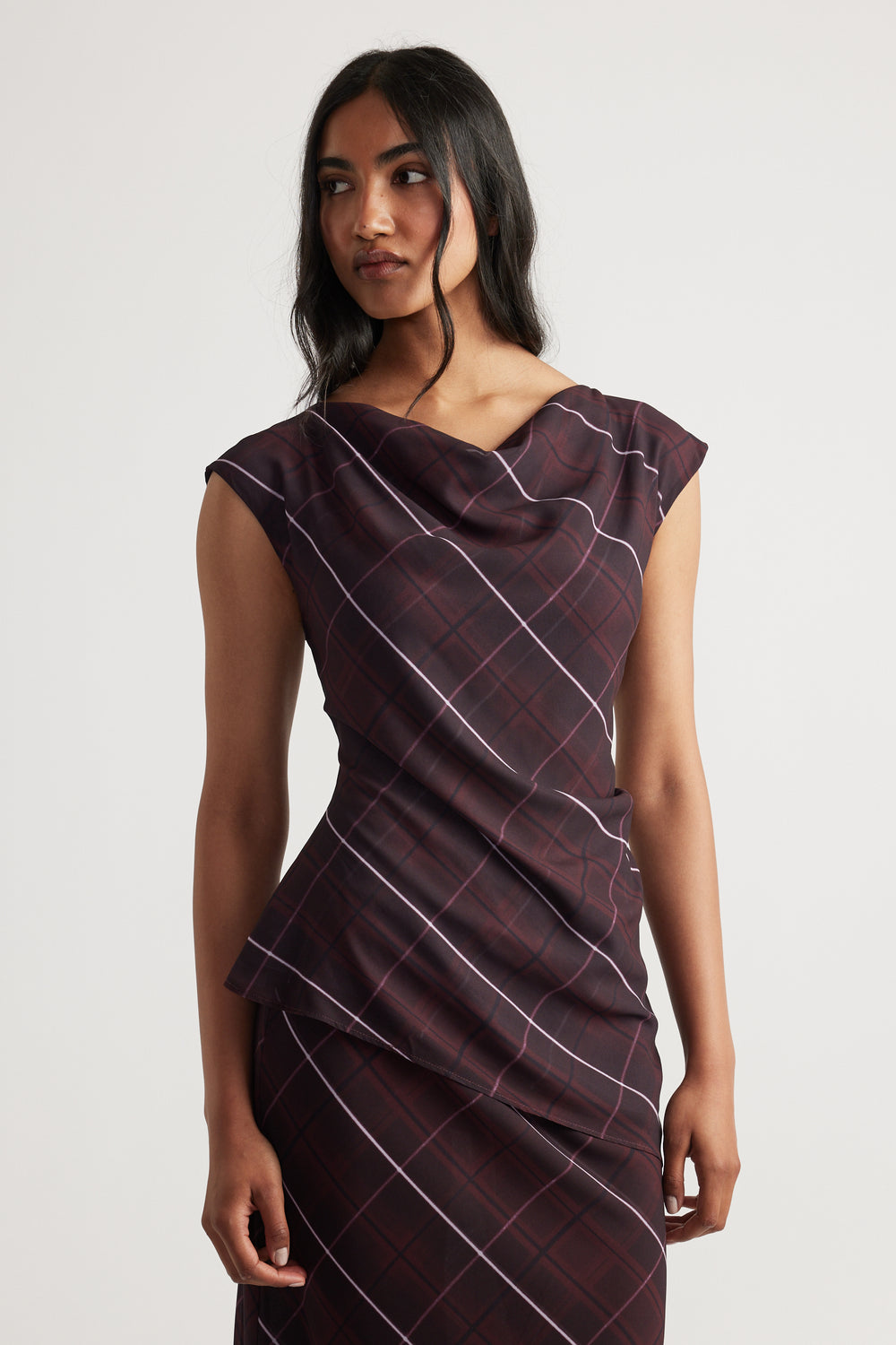 Woman wearing a sleeveless dark plaid top with a draped neckline and matching skirt against a plain backdrop