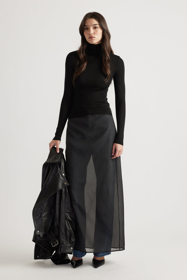 Woman wearing a fitted black knit top paired with layered sheer skirt and denim jeans, holding a black leather jacket.