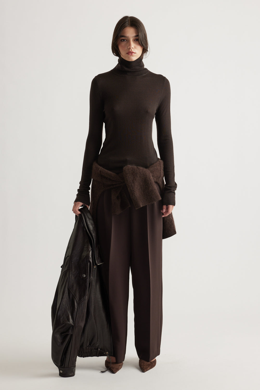 Woman wearing slim-fit chocolate brown wool knit top paired with relaxed dark brown trousers and holding a black jacket