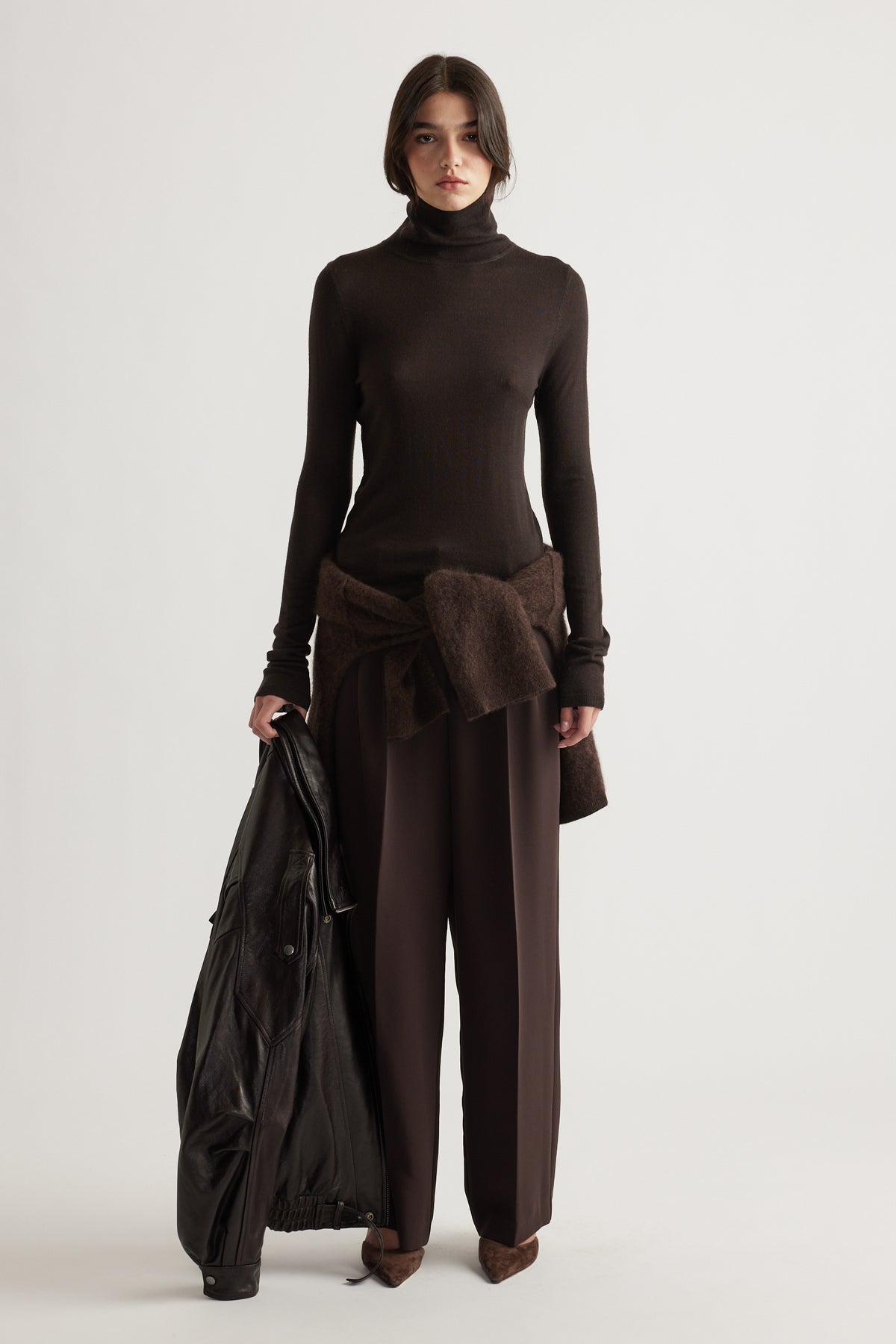 Woman wearing slim-fit chocolate brown wool knit top paired with relaxed dark brown trousers and holding a black jacket