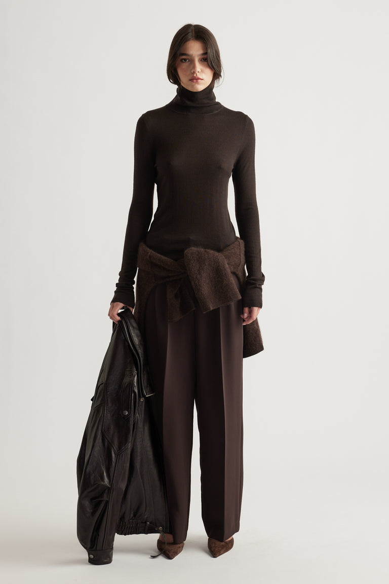 Woman wearing slim-fit chocolate brown wool knit top paired with relaxed dark brown trousers and holding a black jacket