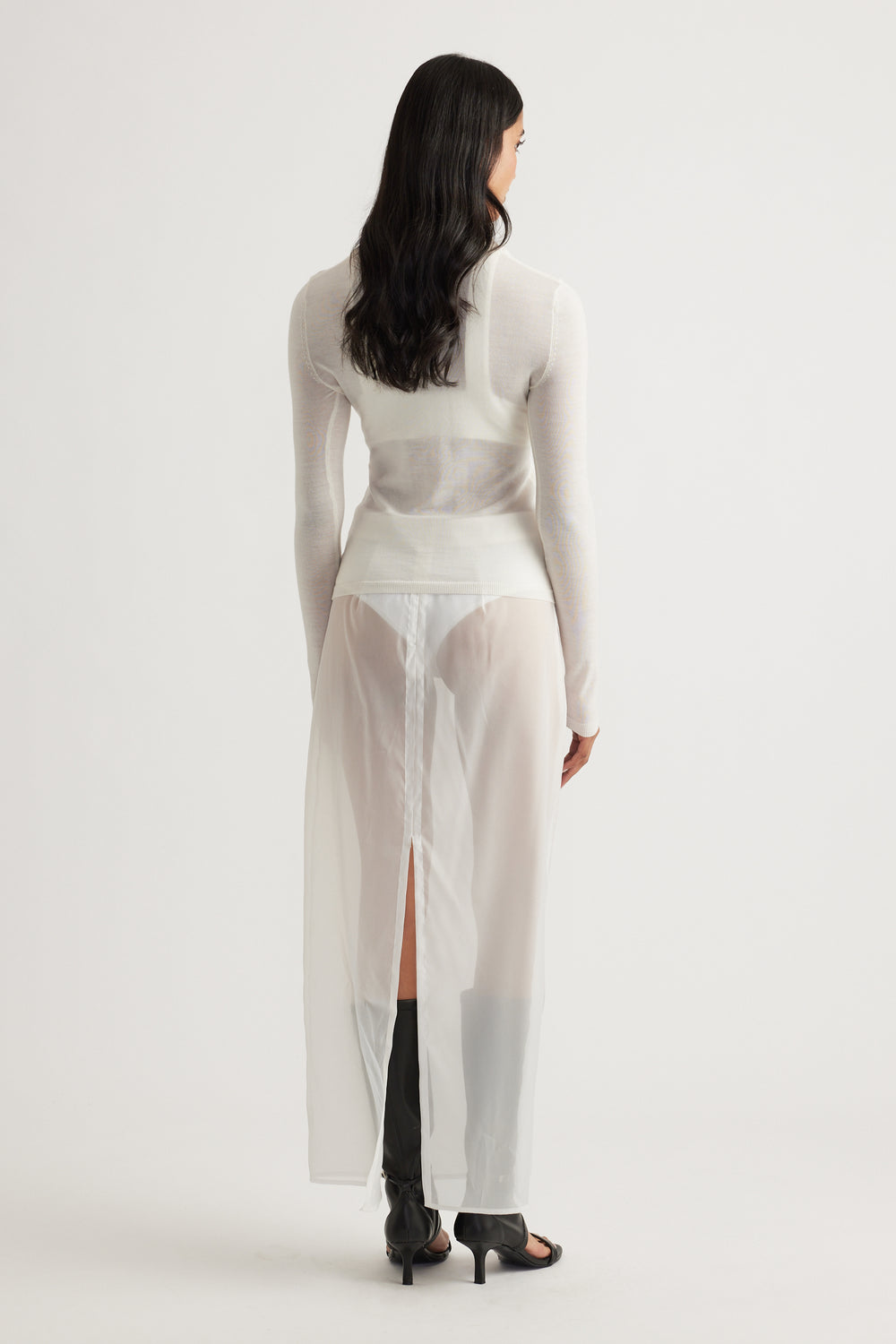 Back view of woman wearing a fitted ivory knit top paired with a sheer long skirt and black heeled boots