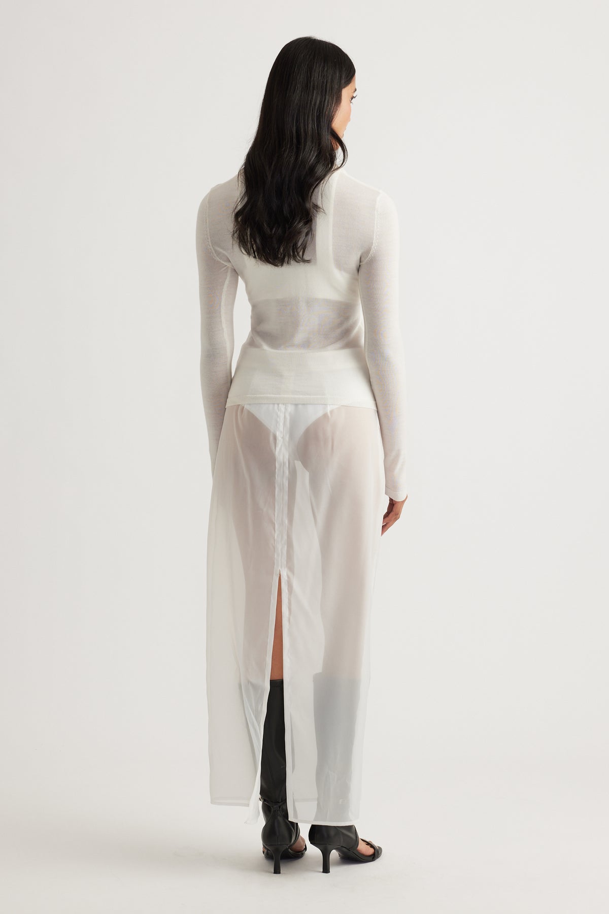 Back view of woman wearing a fitted ivory knit top paired with a sheer long skirt and black heeled boots