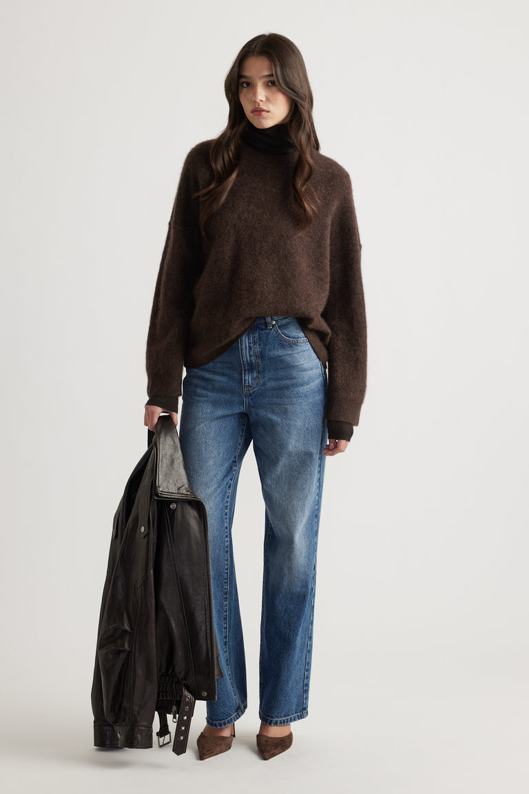 Woman wearing oversized chocolate brown wool knit paired with relaxed blue jeans and holding a black leather jacket
