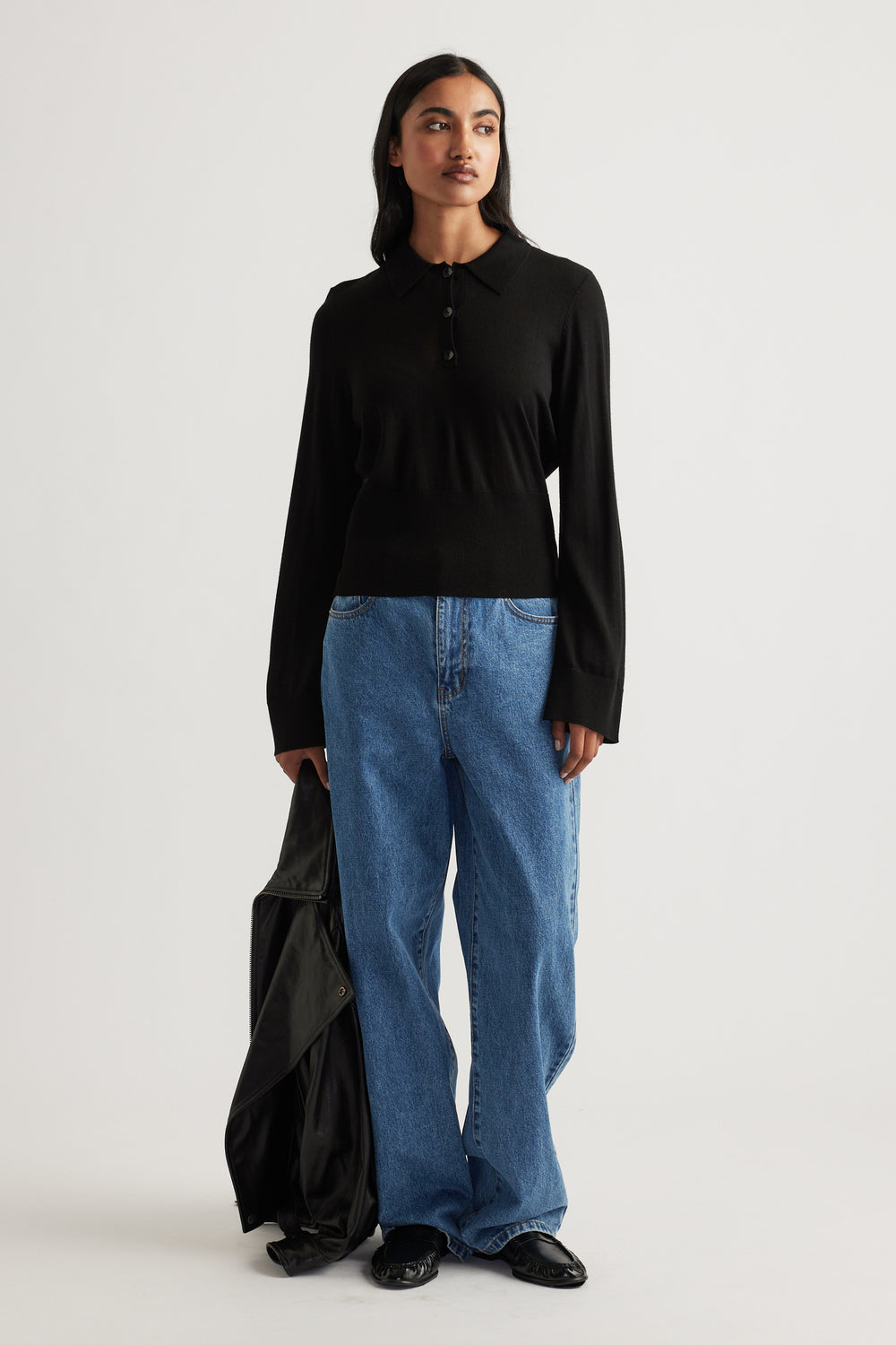 Woman wearing a black fine wool knit polo paired with relaxed blue jeans and holding a black jacket for an effortless look