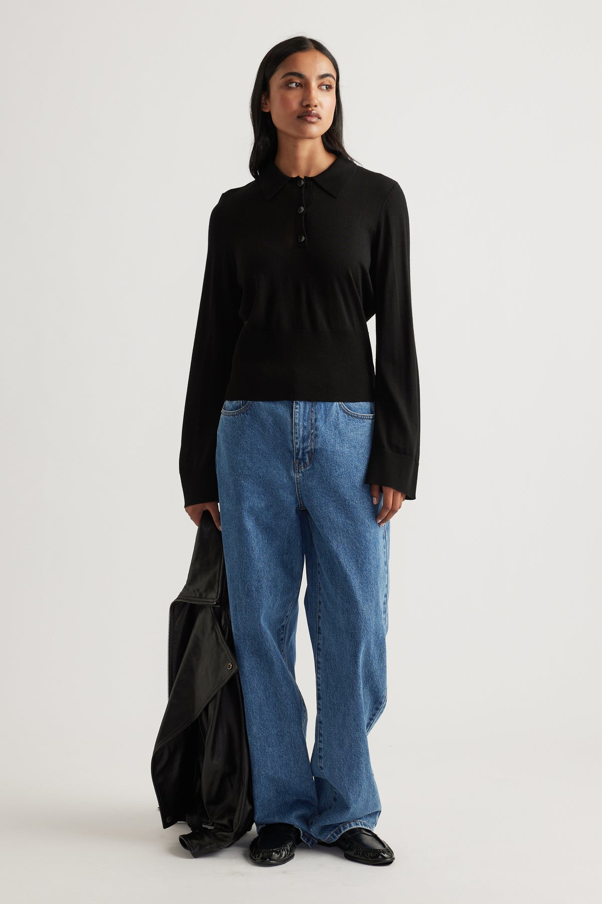 Woman wearing a black fine wool knit polo paired with relaxed blue jeans and holding a black jacket for an effortless look
