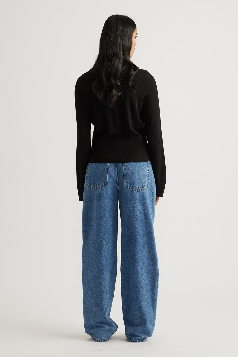 Rear view of woman wearing a fitted black knit top paired with loose straight-leg blue denim jeans against plain background