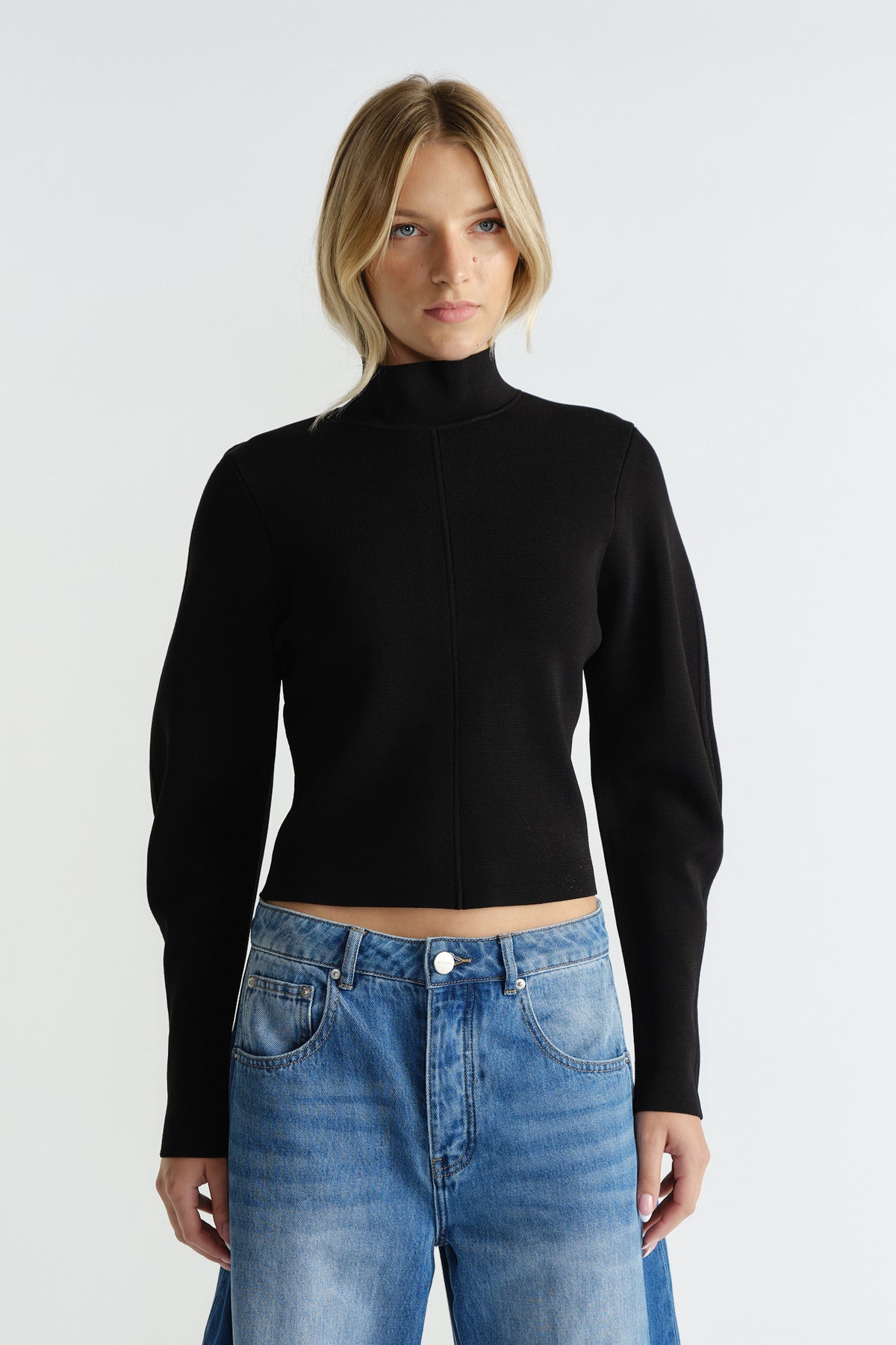 Woman wearing a fitted black high neck jumper paired with relaxed light-wash blue jeans against a neutral background