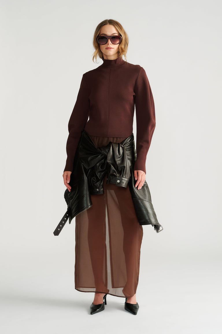 Woman wearing a fitted mocha high neck jumper paired with sheer wide-leg pants and a black leather jacket tied at the waist