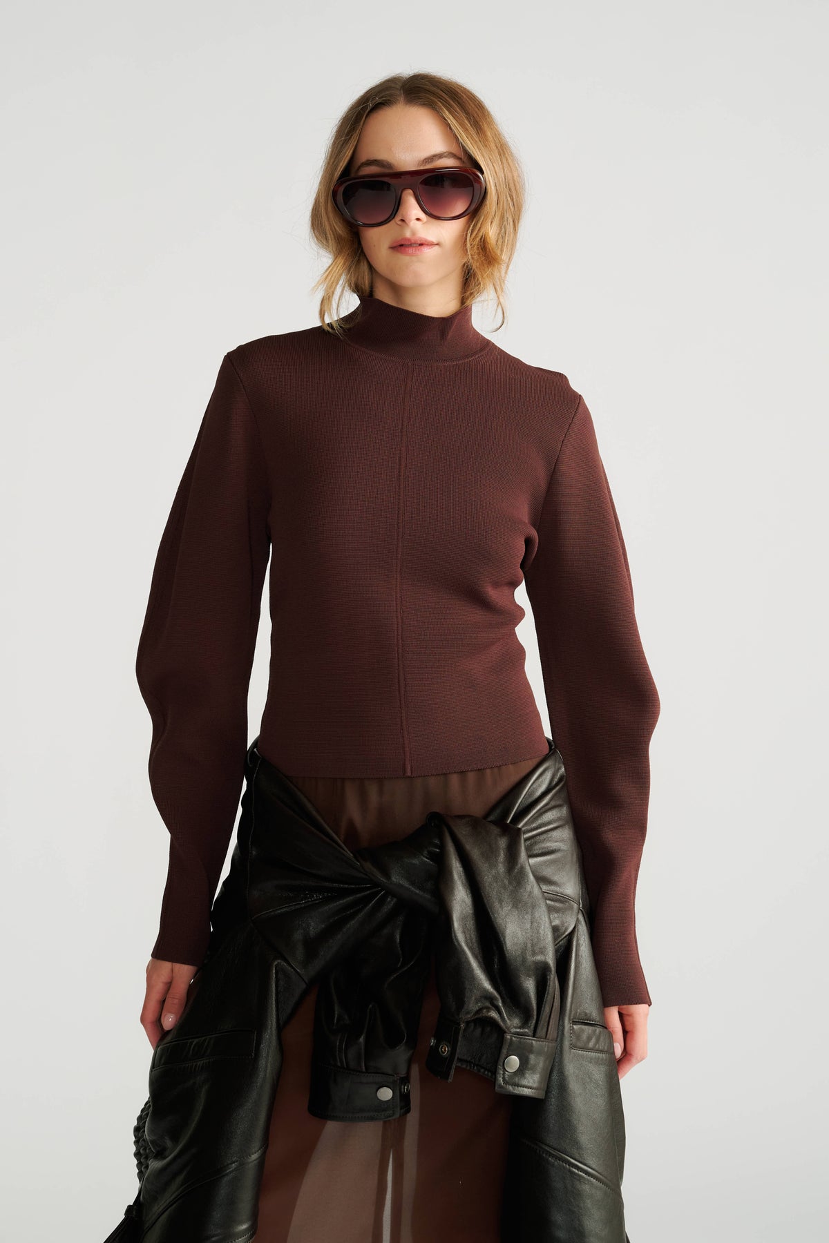 Woman wearing a rich mocha high neck jumper with slim sleeves, paired with a tied black leather jacket at the waist.