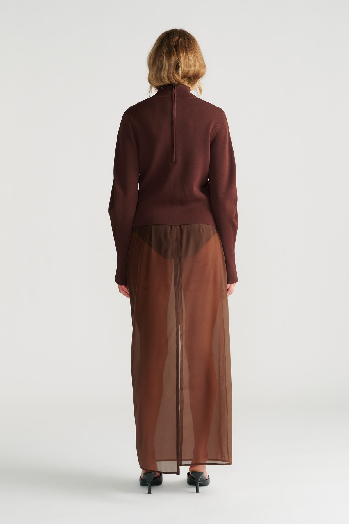 Woman wearing a rich mocha high neck jumper paired with a sheer brown maxi skirt and black heeled sandals, back view.
