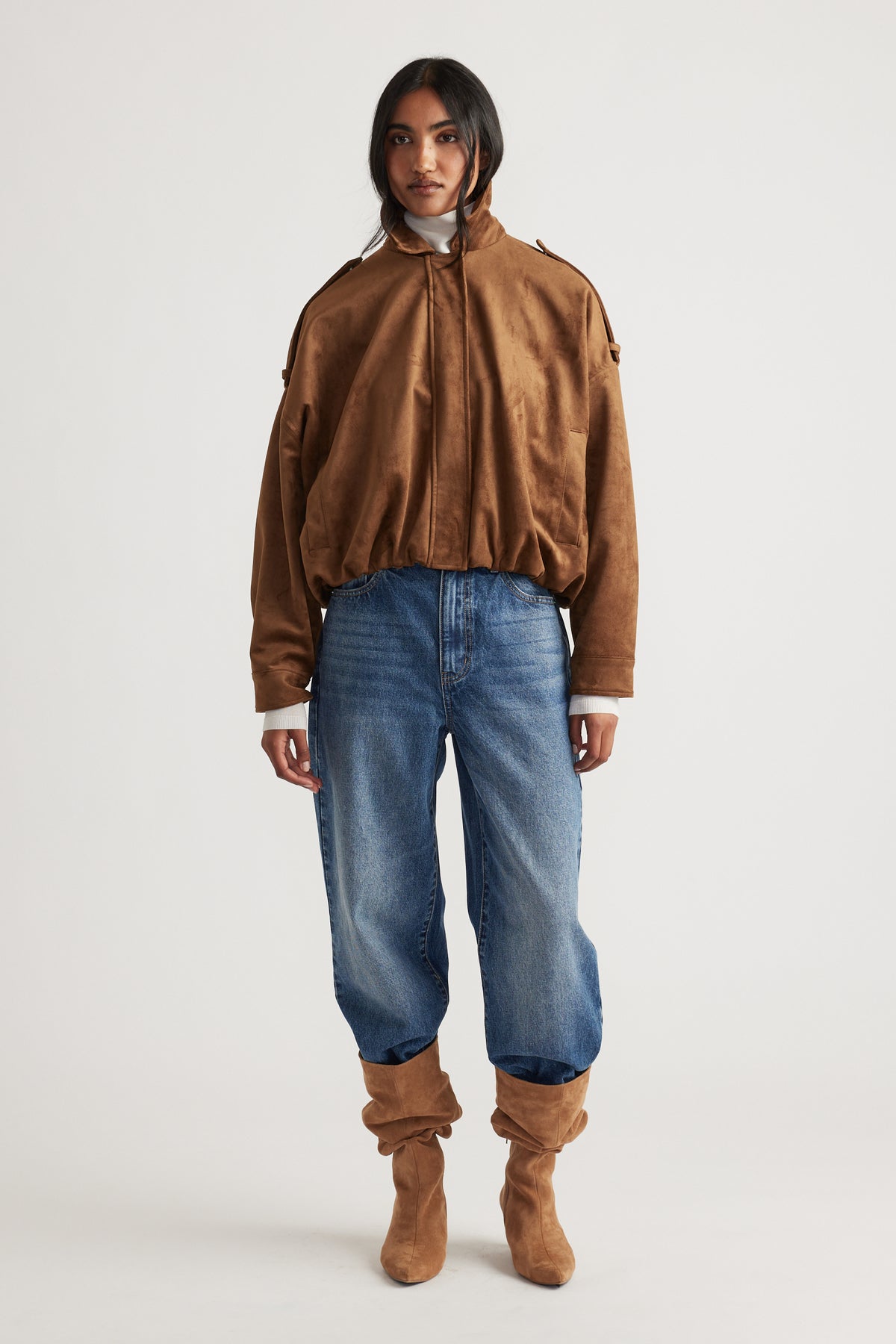 Woman wearing a caramel faux suede bomber jacket with high-waisted relaxed blue jeans and slouchy tan boots