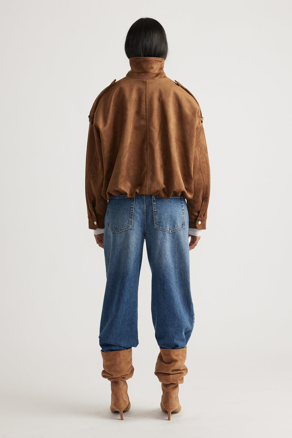 Back view of a caramel faux suede jacket with a high collar, paired with relaxed blue denim jeans and matching boots