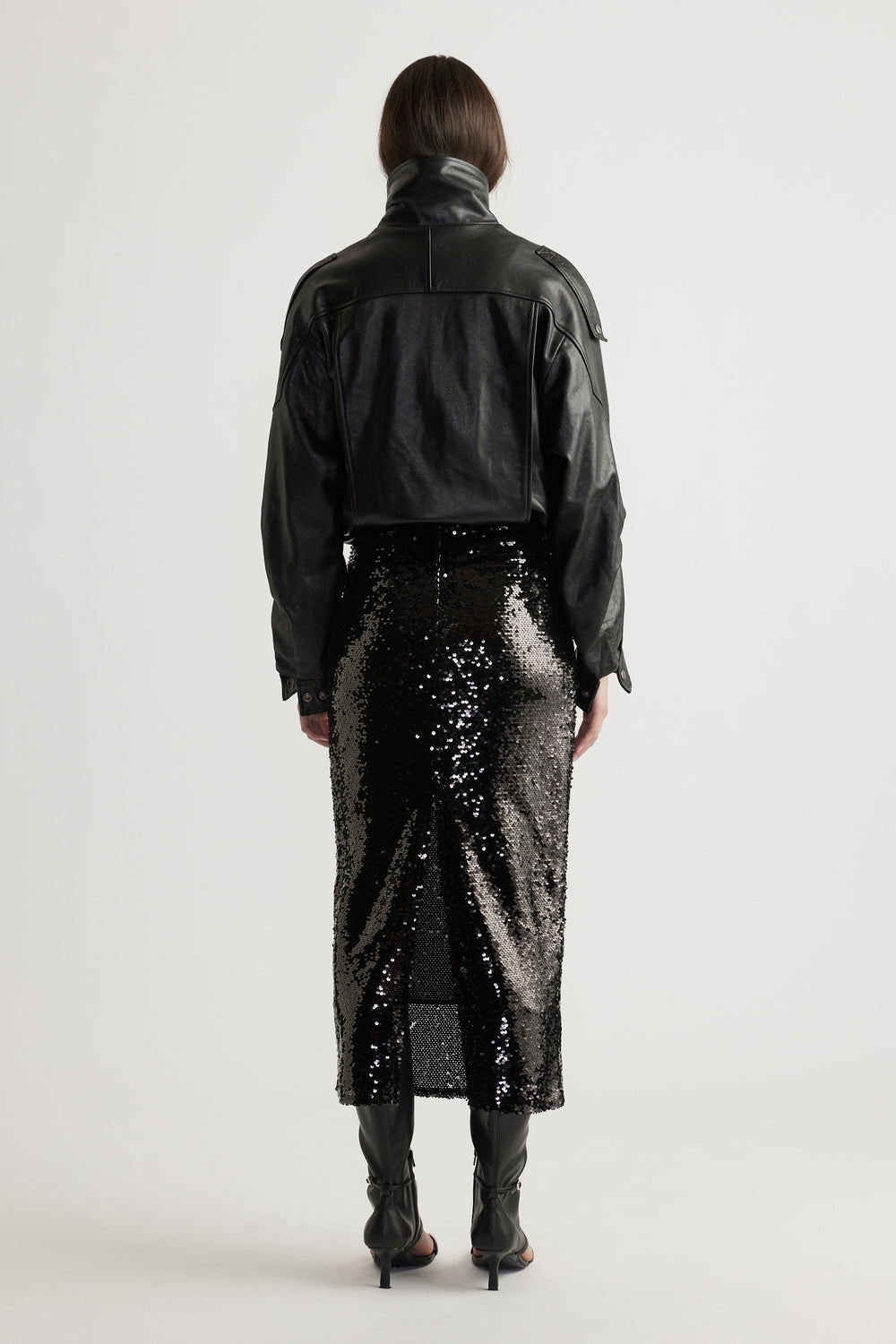 Back view of a woman wearing a black cropped leather jacket paired with a shimmering sequin midi skirt and heeled boots.