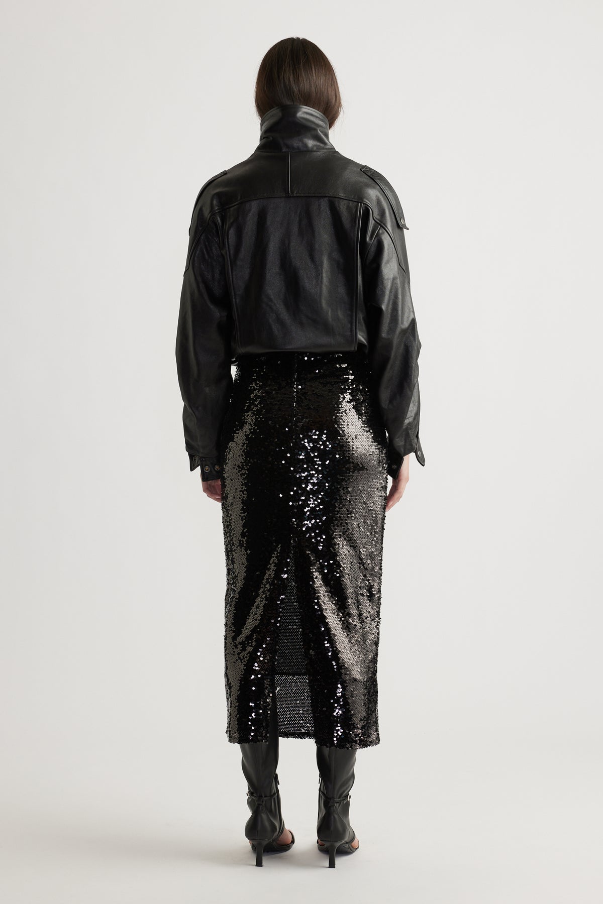 Back view of a woman wearing a black cropped leather jacket paired with a shimmering sequin midi skirt and heeled boots.