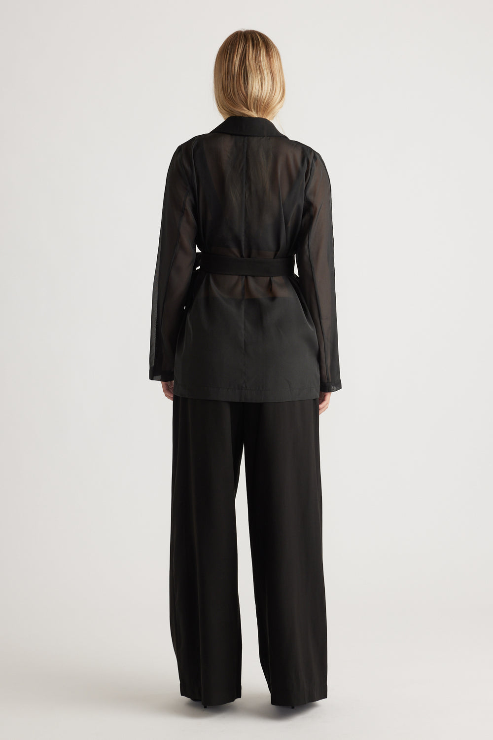 Woman wearing a semi-sheer black blazer with a belted waist paired with wide-leg black trousers, facing away