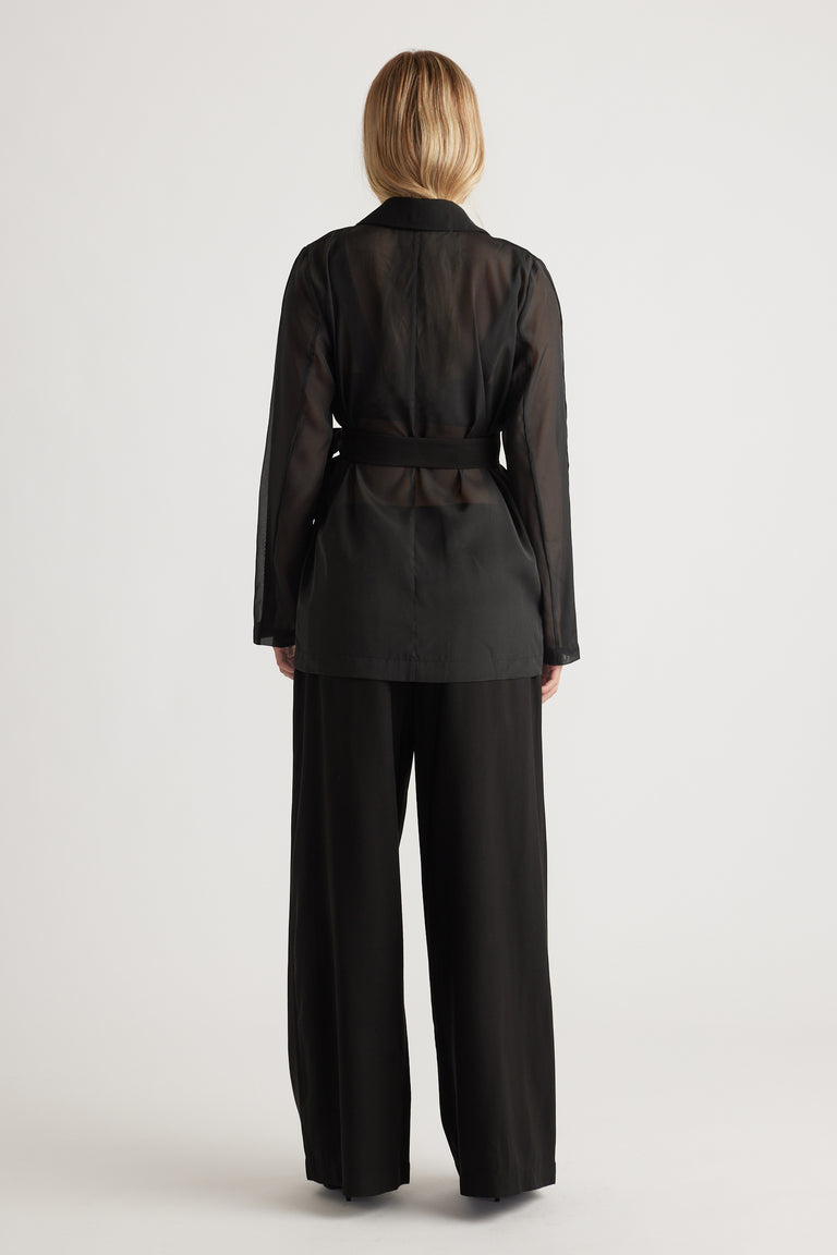 Woman wearing a semi-sheer black blazer with a belted waist paired with wide-leg black trousers, facing away