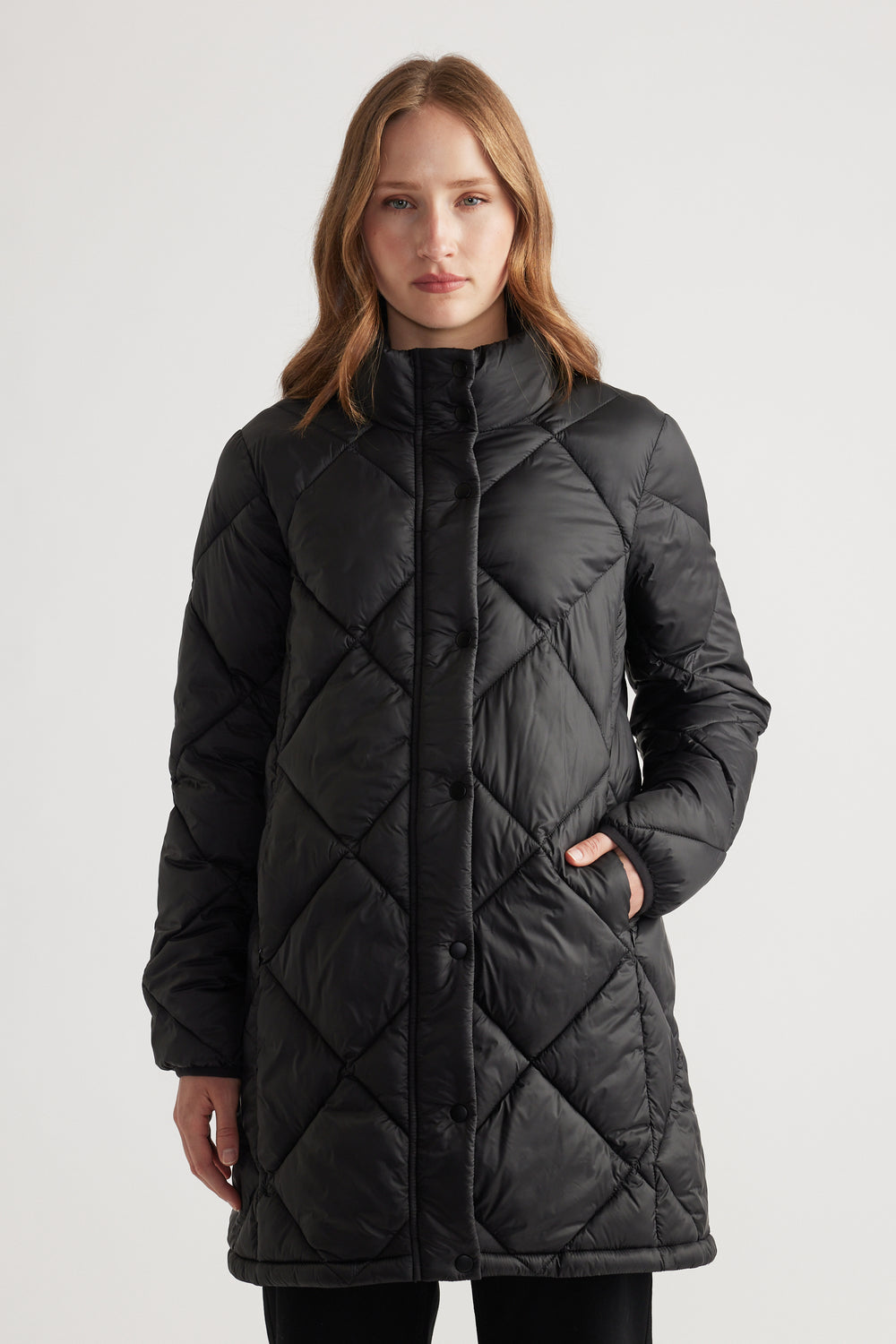 Woman wearing a black quilted longline puffer jacket with high collar and side pockets, standing with a neutral expression