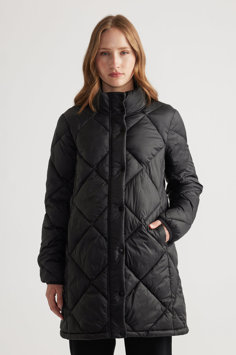 Woman wearing a black quilted longline puffer jacket with high collar and side pockets, standing with a neutral expression