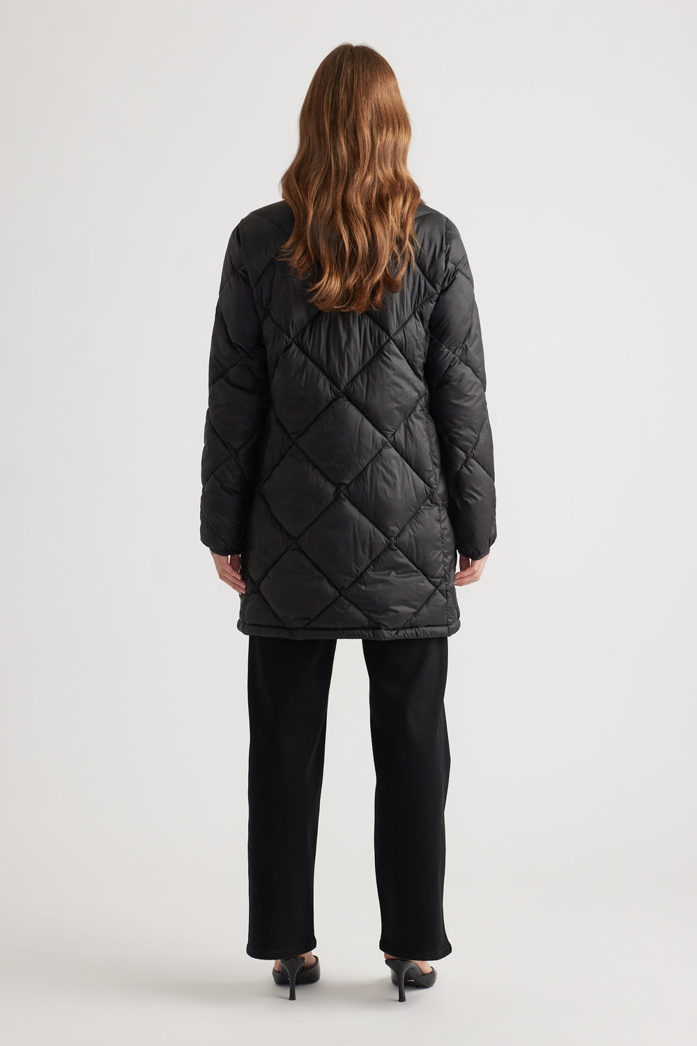 Woman wearing a black quilted longline puffer jacket paired with straight black pants and heels shown from the back