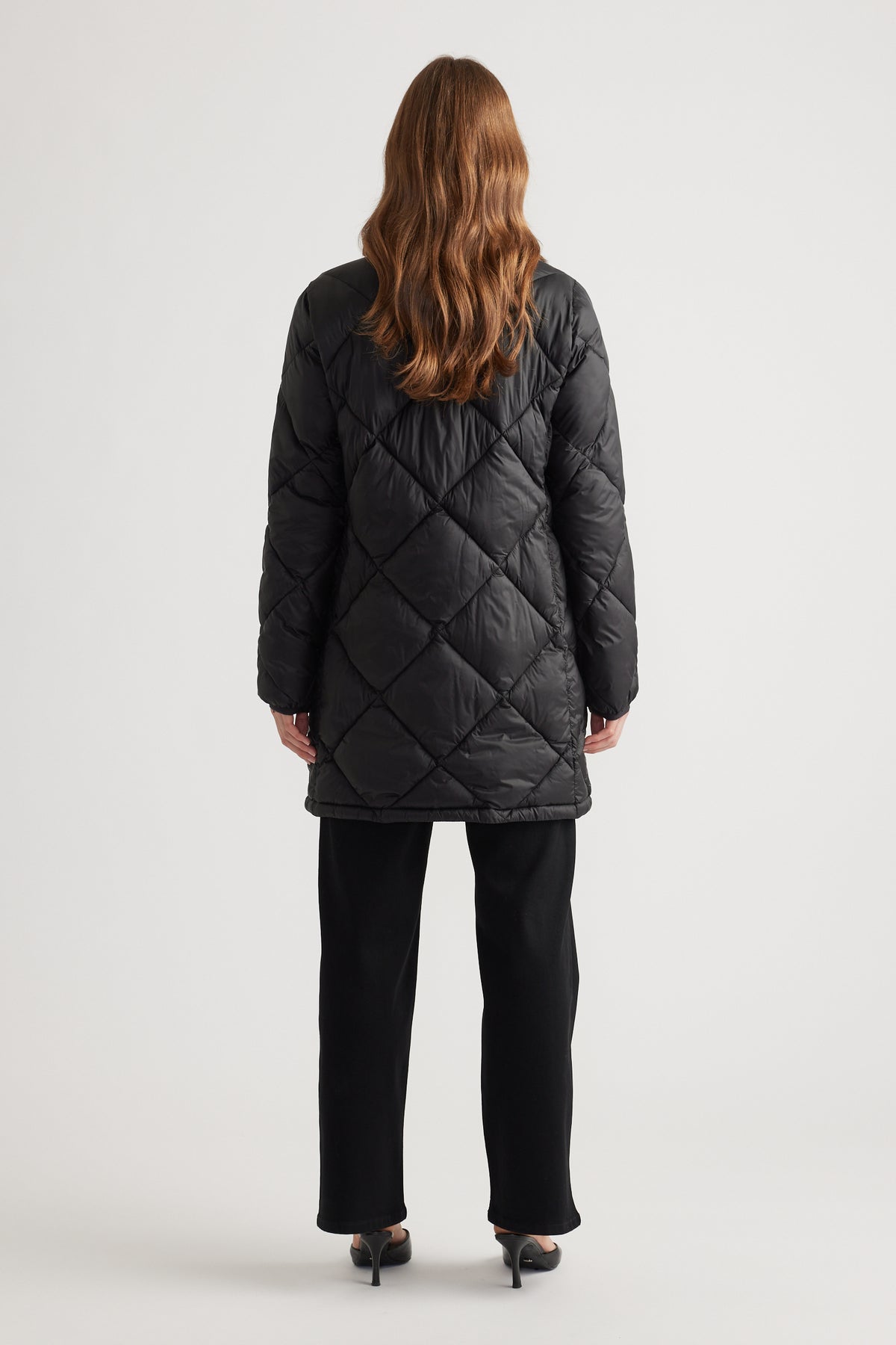 Woman wearing a black quilted longline puffer jacket paired with straight black pants and heels shown from the back