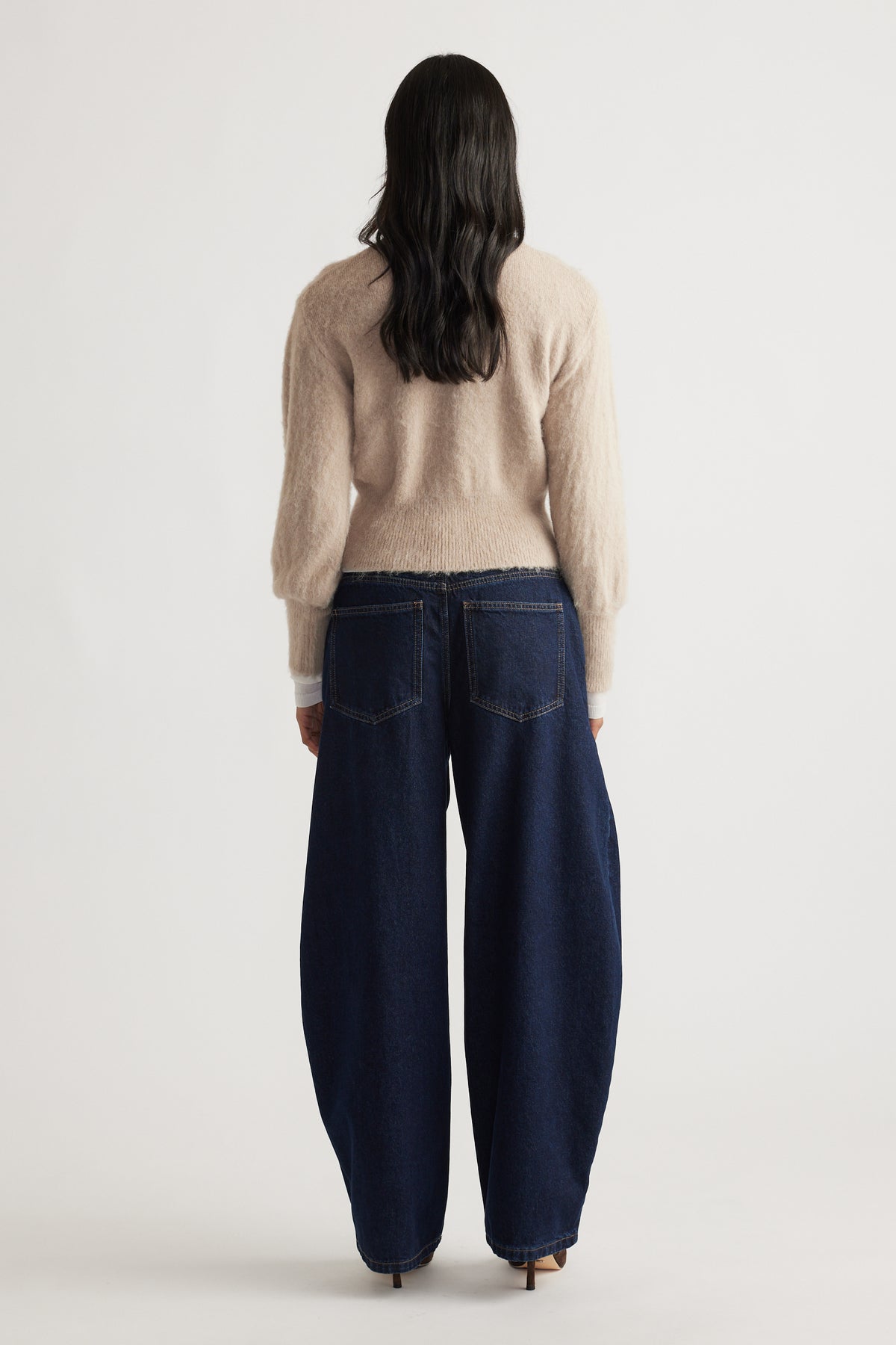 Back view of a woman wearing a soft beige Ariel Button Through Cardigan paired with wide-leg dark denim jeans by DECJUBA