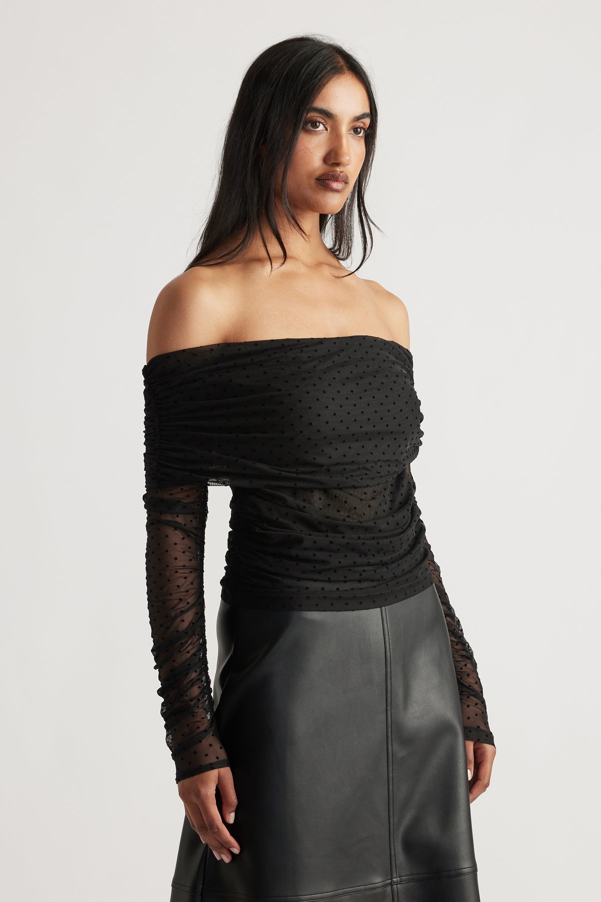 Woman wearing a fitted off-shoulder black mesh top with ruched detailing and long sheer sleeves paired with a leather skirt