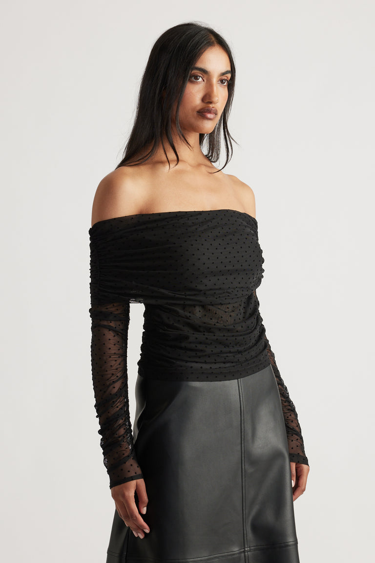 Woman wearing a fitted off-shoulder black mesh top with ruched detailing and long sheer sleeves paired with a leather skirt