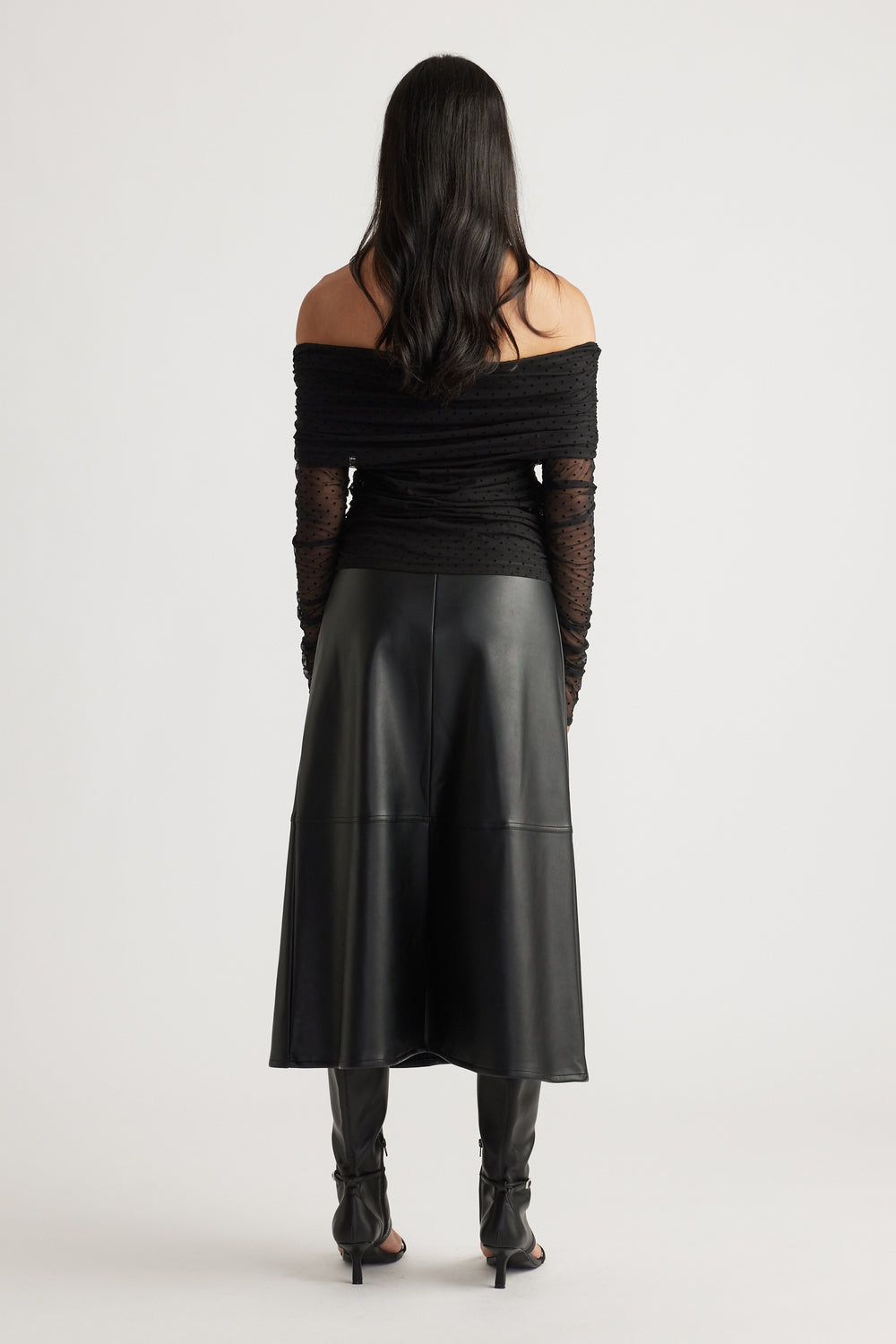 Back view of a woman wearing a fitted off-shoulder ruched mesh top with sheer sleeves, paired with a black midi skirt and heels