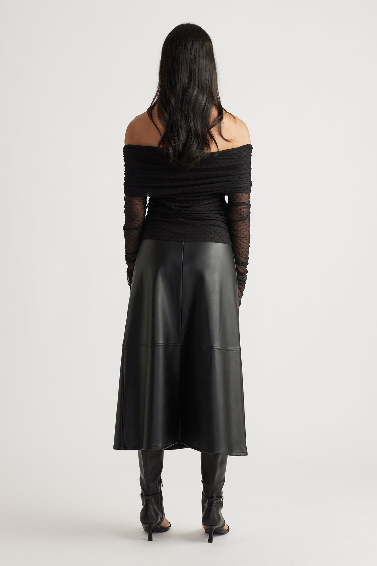 Back view of a woman wearing a fitted off-shoulder ruched mesh top with sheer sleeves, paired with a black midi skirt and heels