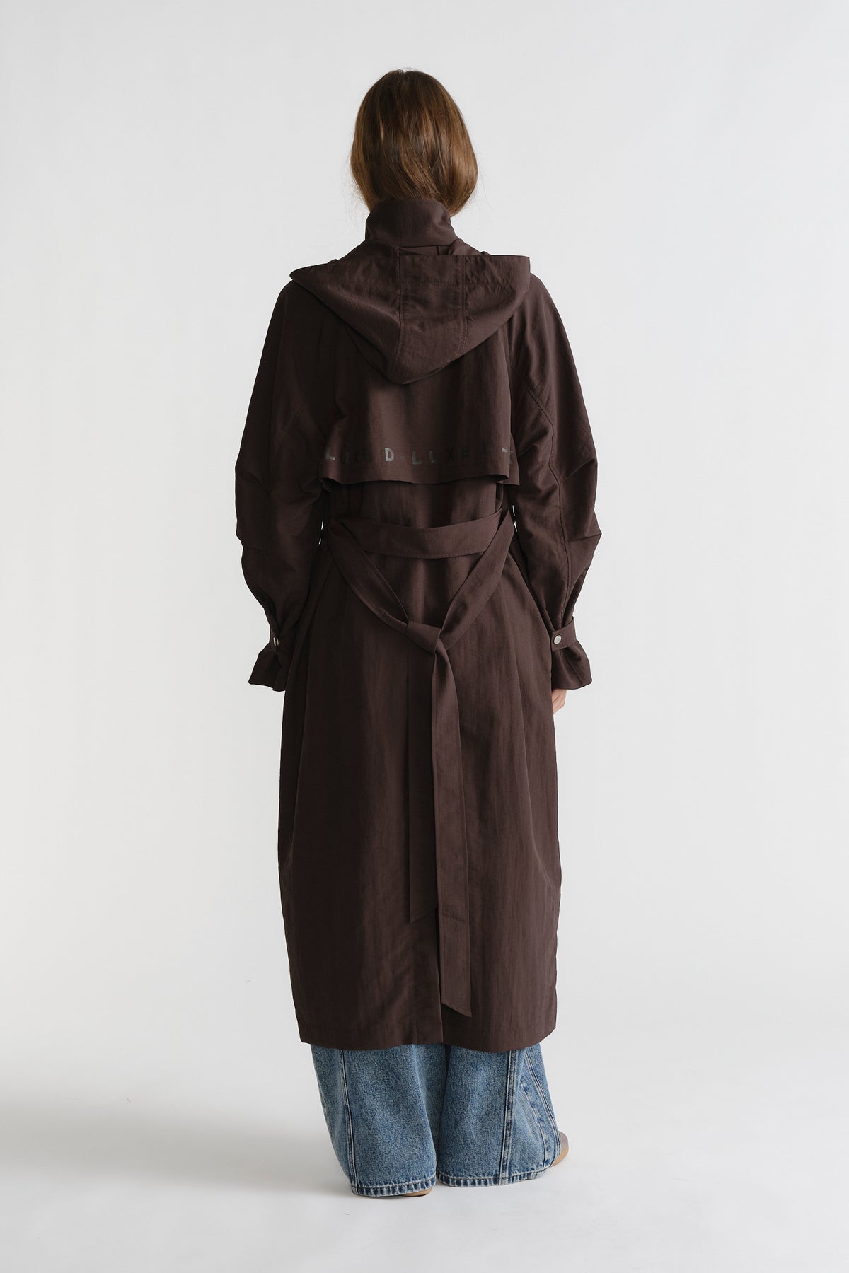 Woman seen from behind wearing cocoa brown D-Luxe Anorak with a belted waist paired with relaxed blue denim jeans.