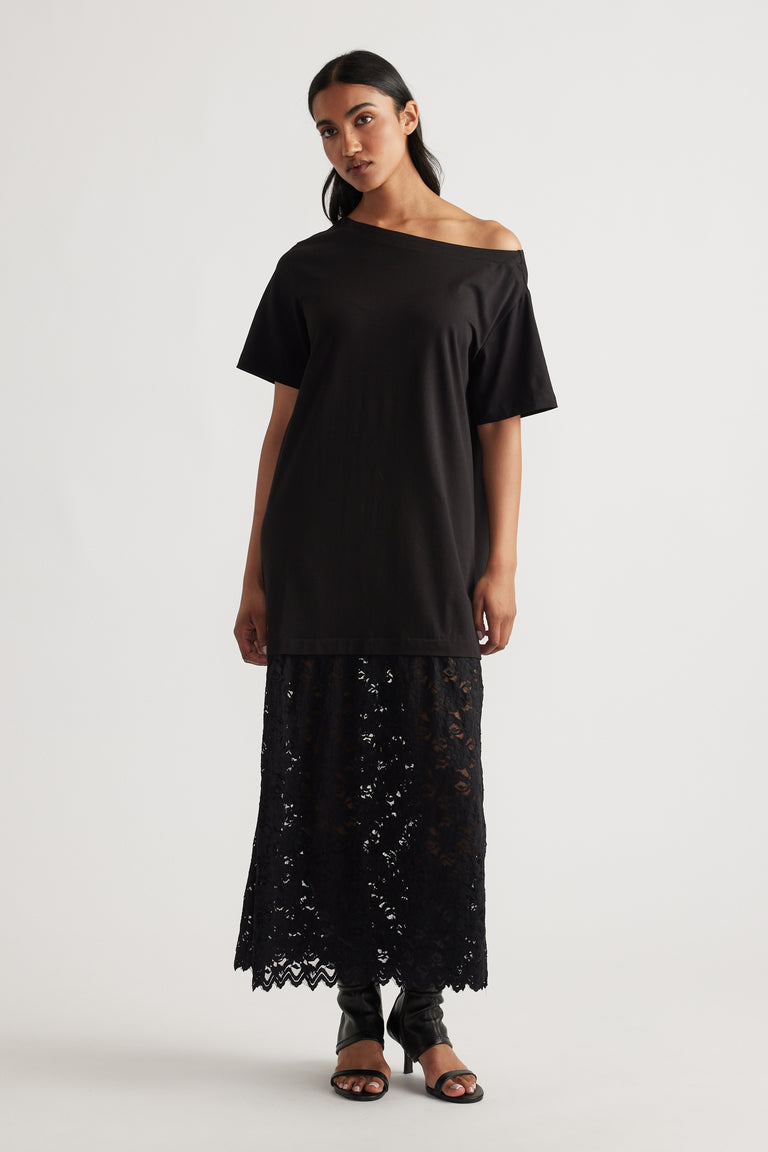 Woman wearing an off-shoulder black top layered over a long black lace skirt, paired with black open-toe heels.