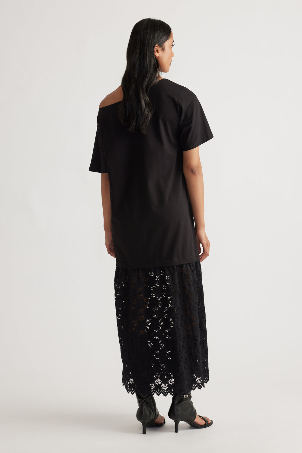 Back view of woman wearing a relaxed black top with an asymmetrical neckline over a sheer black lace maxi skirt and heeled sandals
