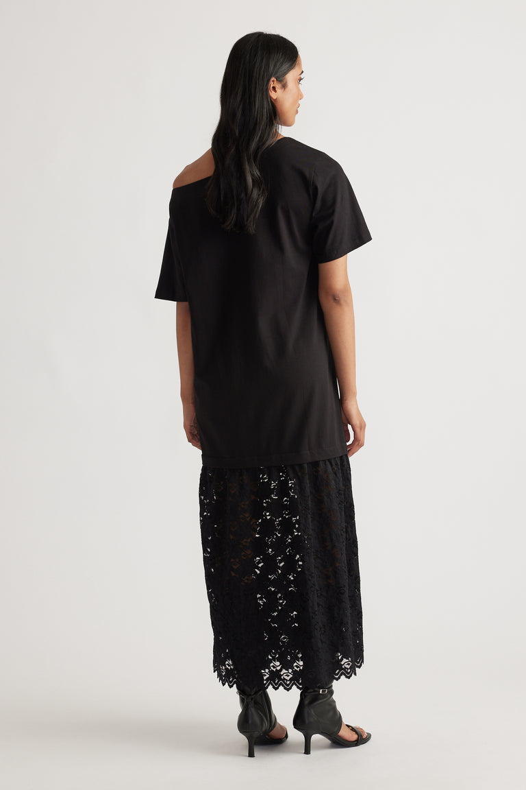 Back view of woman wearing a relaxed black top with an asymmetrical neckline over a sheer black lace maxi skirt and heeled sandals