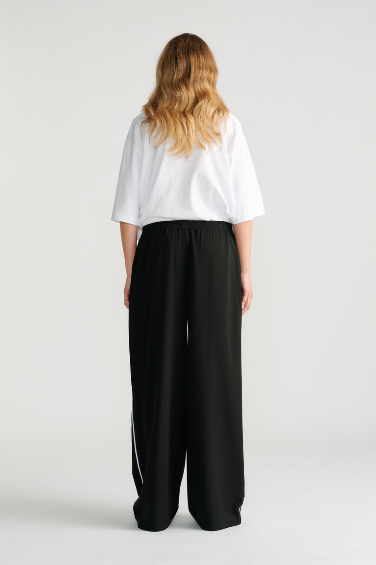 Woman wearing black wide-leg twill pants with a white side stripe paired with a loose white short-sleeve top.