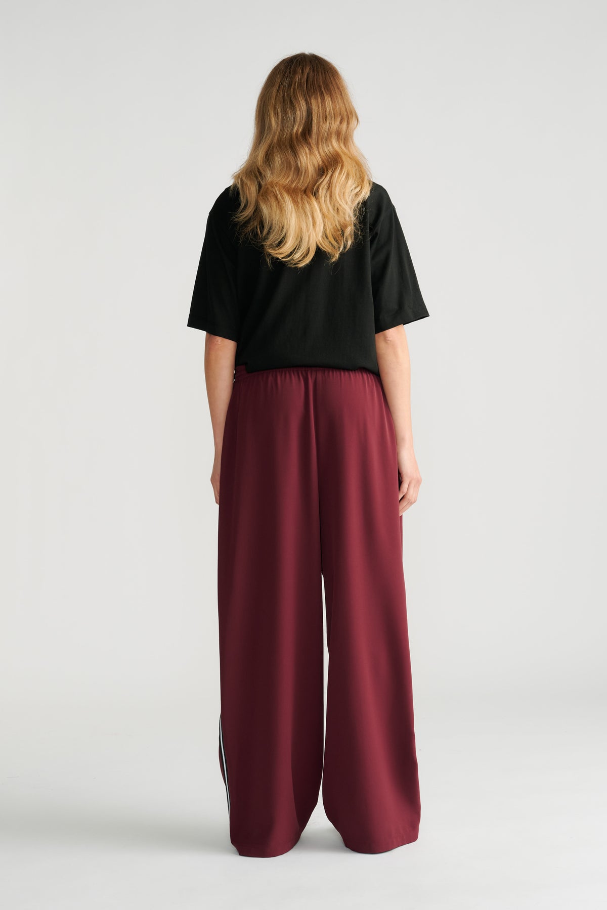 Woman wearing relaxed plum twill pants with side stripes, styled with a loose black short-sleeve top, shown from behind.