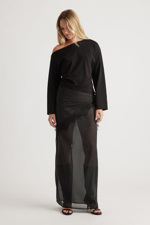 Woman wearing an off-shoulder black long sleeve top paired with a sheer black maxi skirt featuring lace trim details