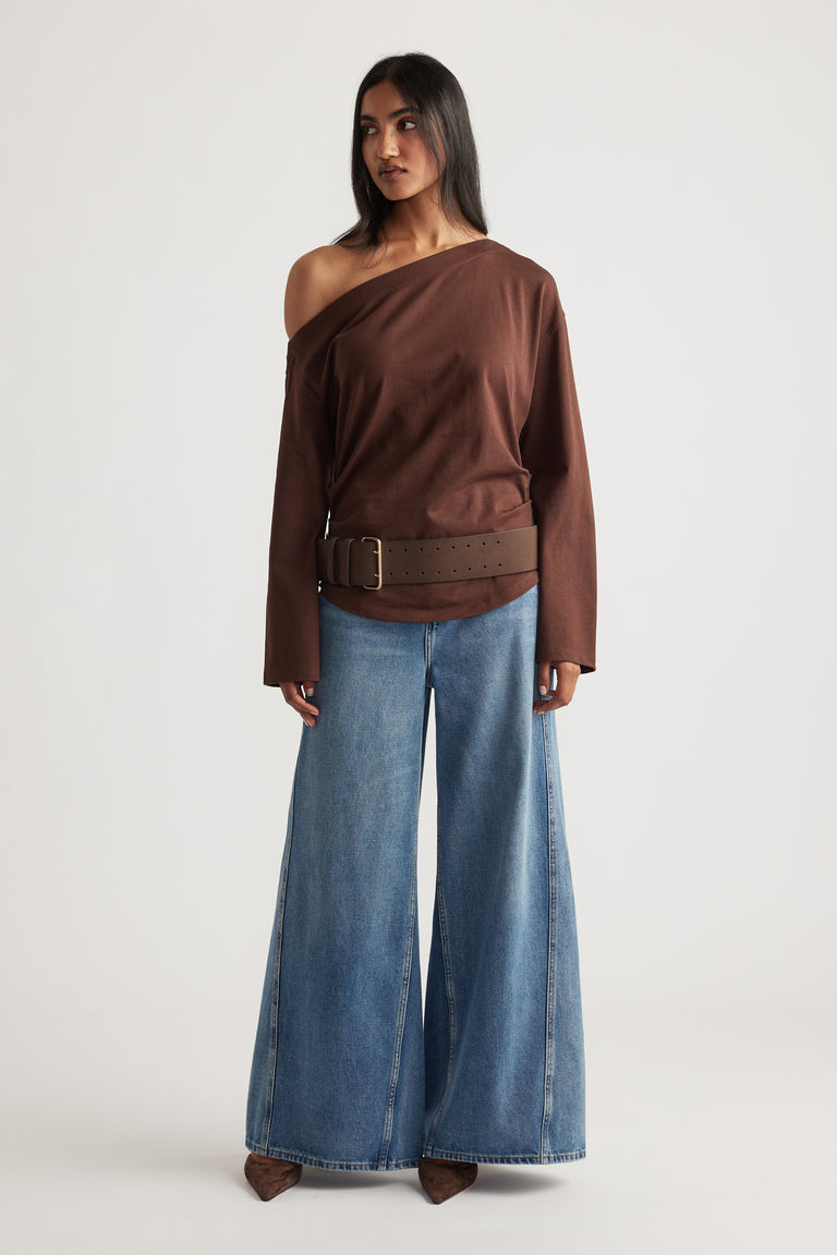 Woman wearing an off-shoulder brown top with long sleeves and a wide belt, paired with wide-leg blue denim jeans