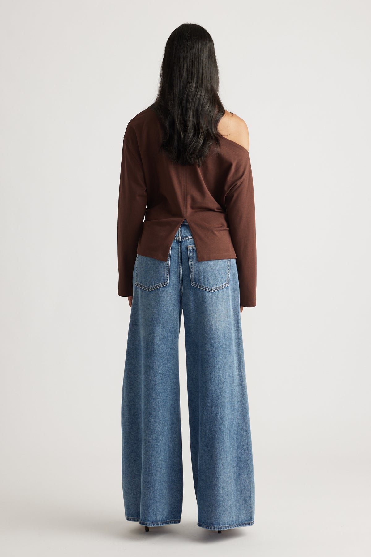 Back view of a woman wearing a brown off-shoulder top with a back slit, paired with wide-leg blue denim jeans