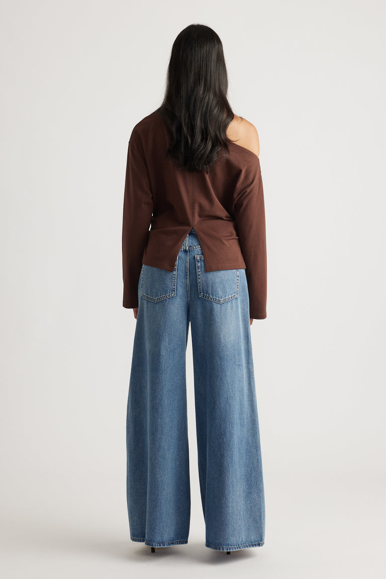 Back view of a woman wearing a brown off-shoulder top with a back slit, paired with wide-leg blue denim jeans