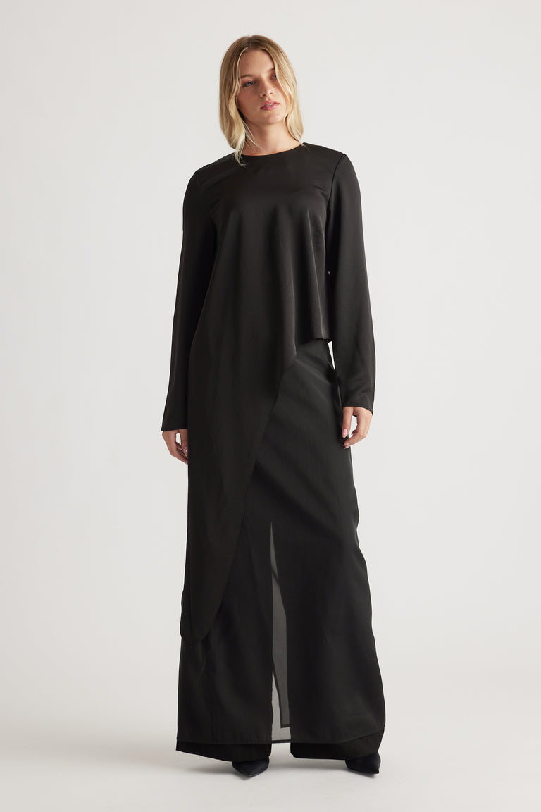 Front view of a woman wearing a black long-sleeve draped top with wide-leg black pants and pointed shoes