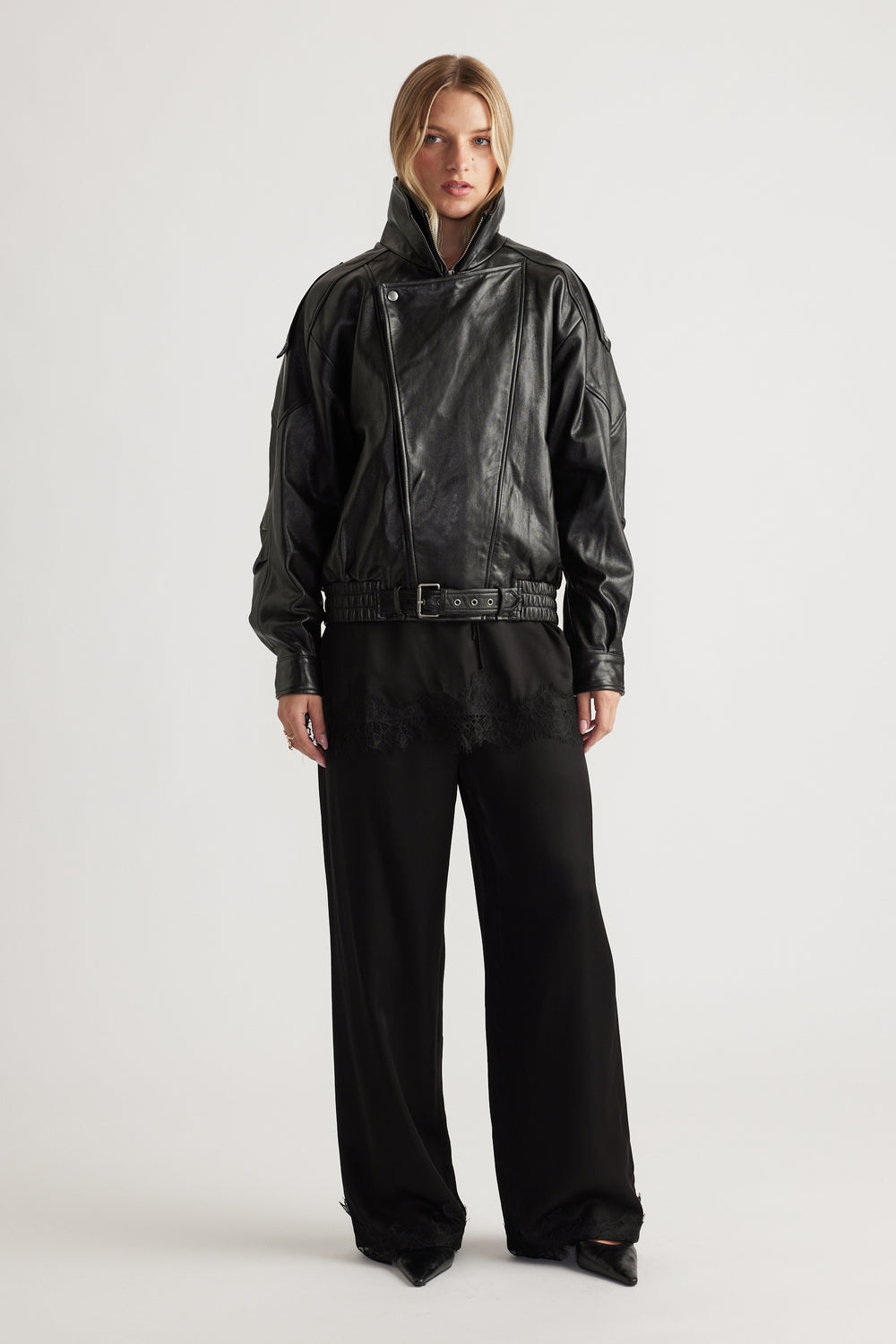 Woman wearing a black belted leather jacket styled with loose black satin pants featuring delicate lace trim at the hem