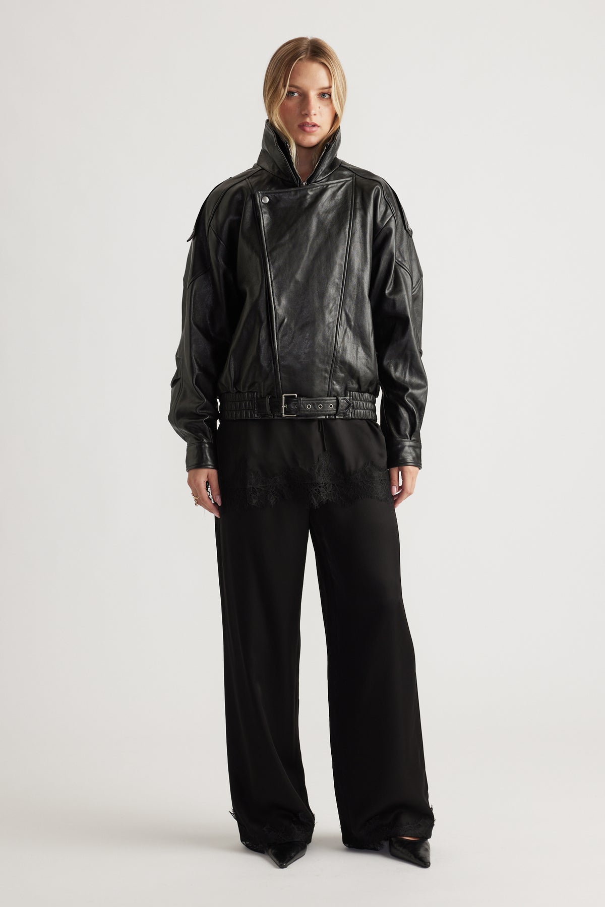 Woman wearing a black belted leather jacket styled with loose black satin pants featuring delicate lace trim at the hem