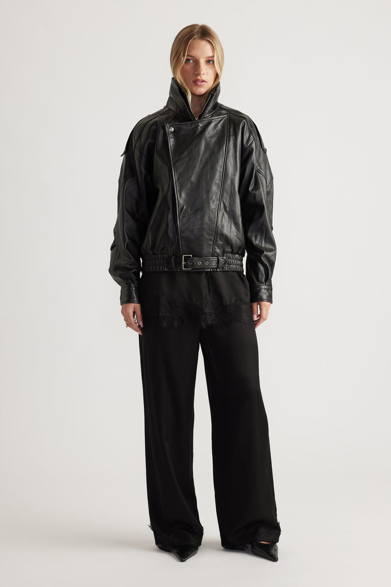 Woman wearing a black belted leather jacket styled with loose black satin pants featuring delicate lace trim at the hem